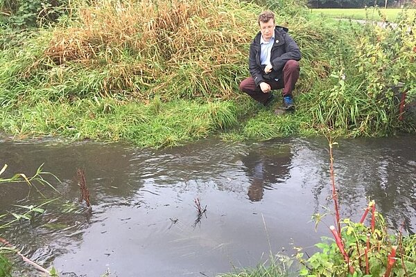 Cllr Will Frass kneeling beside a flood Fairywell Brook Cllr Will Frass kneeling beside a flooded Fairywell Brook